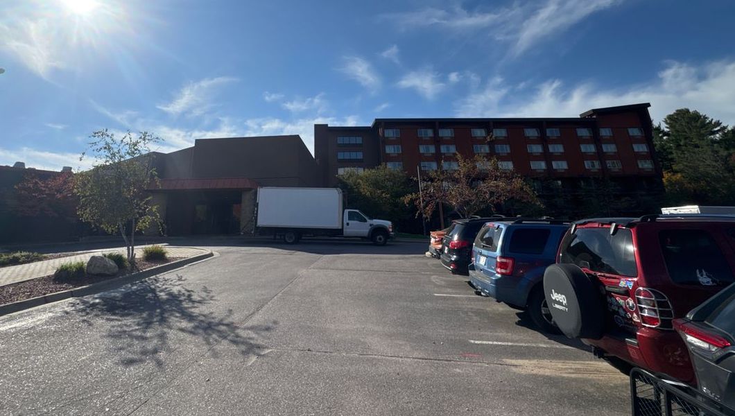The building from the parking lot: a low brown entrance with a reddish awning on the left, and the taller red-brown hotel wing rising behind it on the right.
